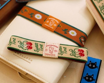 May include: A collection of elastic bands for notebooks featuring various designs. One band is beige with red cherries and the text Sweet Time, another is orange with white daisies, and a third is blue with black cat faces. Each has a central label.