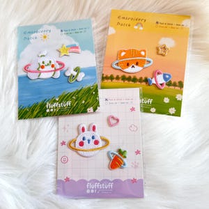 Op de afbeelding: Drie verpakkingen met geborduurde patches met cartoonontwerpen. Eén verpakking heeft een wit konijn met een planeetring, een wortel en een roze hart. Een andere verpakking heeft een kat met een planeetring en een raket. De derde verpakking heeft een wolk met een planeetring en een ster.
