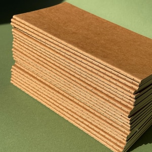 May include: A stack of rectangular notebooks with plain brown paper covers. Each notebook features a visible stitched spine along the left edge, showing the thread binding that holds the pages together in a slim, vertical orientation.