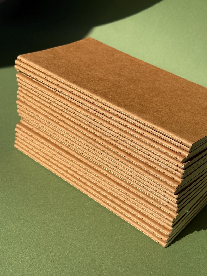 May include: A stack of rectangular notebooks with plain brown paper covers. Each notebook features a visible stitched spine along the left edge, showing the thread binding that holds the pages together in a slim, vertical orientation.