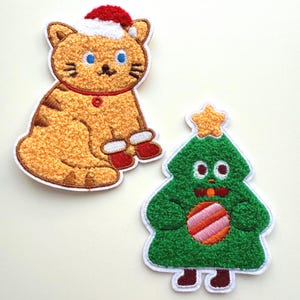 May include: Two embroidered patches: a cat wearing a Santa hat and mittens, and a Christmas tree with a star on top. The cat is orange with brown stripes, blue eyes, and red accents. The tree is green with a striped ornament.