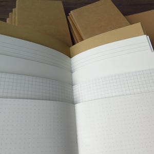 May include: A collection of notebooks with plain brown kraft paper covers. The notebooks are open to reveal various interior page styles, including white pages with horizontal lines, white pages with a grid pattern, and white pages with a dot grid.