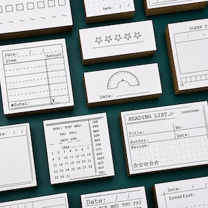 Könnte beinhalten: Eine Sammlung rechteckiger Holzstempel mit weißen Oberseiten und schwarzen Tintendesigns für Journaling und Planung. Die Stempel enthalten Vorlagen für Leselisten, Schlaf-Tracking, Kalender, Ausgabenprotokolle und Tagespläne mit verschiedenen Symbolen.