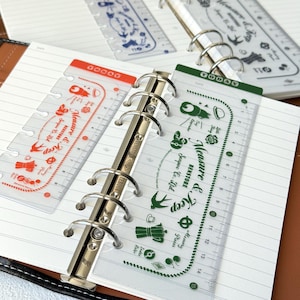 May include: Clear plastic index rulers for ring-bound planners, featuring printed graphics of coffee pots, swallows, and the text Measure & Keep. The rulers include a 15-centimetre scale and a Today tab, available in orange and green ink colour options.