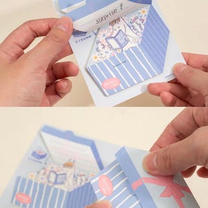 May include: A light blue and white striped paper gift box with a pink bow. The box is being opened to reveal a cartoon illustration of a character reading a book. The word "surprise!" is written on the inside flap.