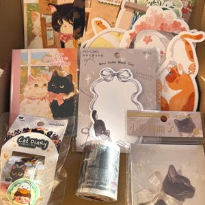 May include: A collection of cat-themed stationery items in a cardboard box. Includes stickers, washi tape, notepads, and cards featuring illustrations of cats. The items have various designs, including cartoon cats and floral elements. Some items have text like "Cat Diary" and "Bow Little Black Cat".