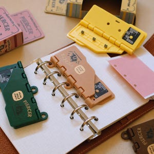 May include: A collection of plastic mini hole punch tools in yellow, green, peach, and brown, each labeled Piercer and Mini Hole Punch. These tools are designed to snap into the metal rings of a grid-lined notebook, with some shown attached to the paper.