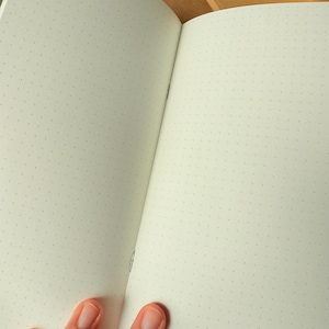 May include: An open notebook with off-white pages featuring a light gray dot grid pattern. The notebook has a simple stitched binding visible at the center fold, and the paper has a smooth texture. Additional closed brown paper notebooks are visible.