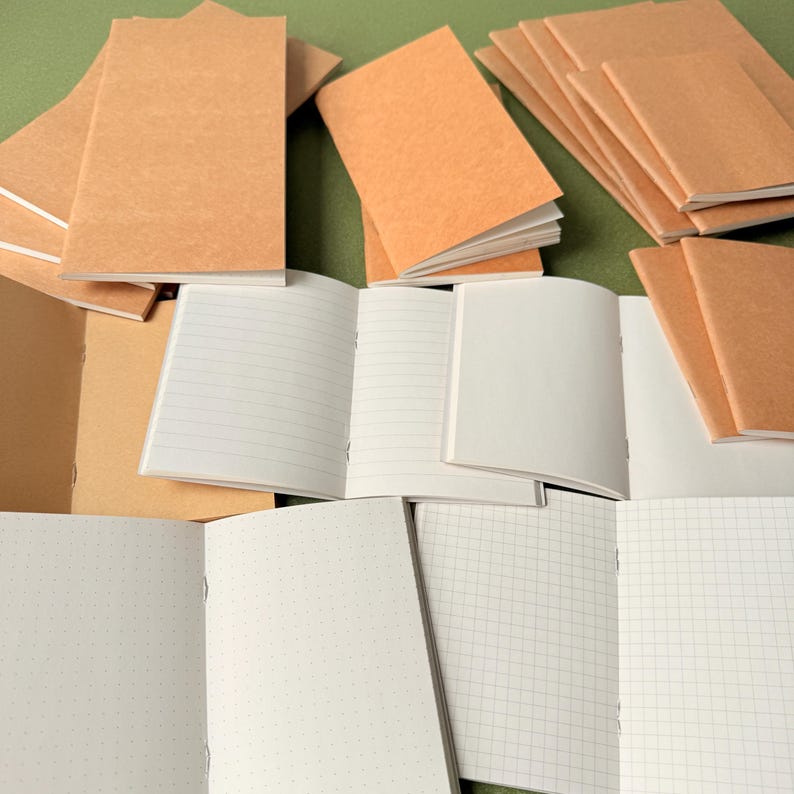 May include: A collection of small notebooks with light brown paper covers. Some notebooks are closed and stacked, while others are open to reveal white interior pages featuring various layouts including horizontal lines, dot grids, and square grids.