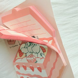 May include: A stack of pink memo pads with a strawberry cake design. The cake features layers of pink frosting, strawberries, and a cartoon illustration of Moomin characters. The memo pads are in a pink paper holder.
