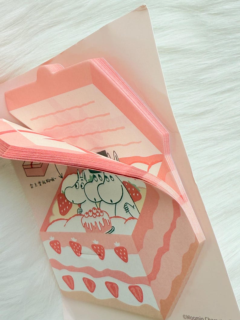May include: A stack of pink memo pads with a strawberry cake design. The cake features layers of pink frosting, strawberries, and a cartoon illustration of Moomin characters. The memo pads are in a pink paper holder.
