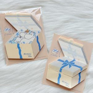 May include: Two beige gift box-shaped notepads with blue ribbon designs. One notepad is open, revealing a cartoon character and stars. The other is closed. The text "Moomins Letters" is visible on both.