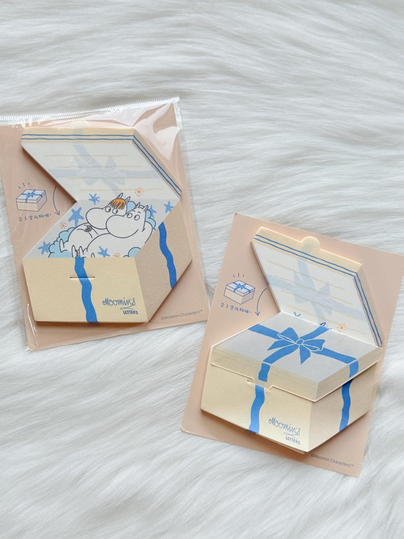 May include: Two beige gift box-shaped notepads with blue ribbon designs. One notepad is open, revealing a cartoon character and stars. The other is closed. The text "Moomins Letters" is visible on both.