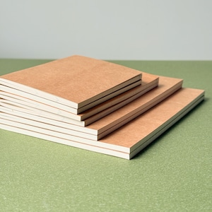 May include: A stack of four square notebooks with plain, light brown cardboard covers. Each notebook features a thick, tan-colored paper cover and a visible white paper edge, arranged in a descending size order from top to bottom.
