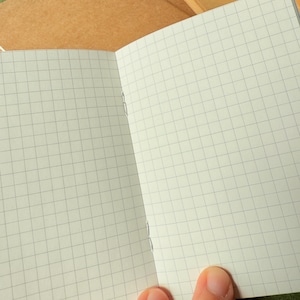 May include: An open notebook with light cream-colored pages featuring a thin gray grid pattern. The notebook is bound with visible silver staples along the center fold. Additional closed notebooks with plain brown covers are visible in the background.