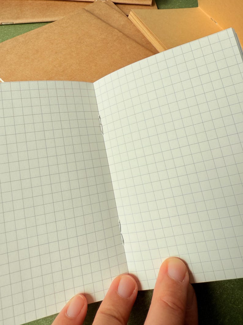 May include: An open notebook with light cream-colored pages featuring a thin gray grid pattern. The notebook is bound with visible silver staples along the center fold. Additional closed notebooks with plain brown covers are visible in the background.