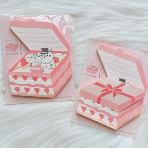 May include: Two pink and white notepad sets shaped like gift boxes. One shows a cake with strawberries and Moomin characters. The other has a pink bow on top. Each notepad has a hinged lid.