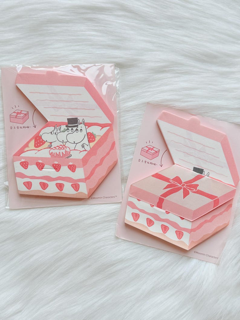May include: Two pink and white notepad sets shaped like gift boxes. One shows a cake with strawberries and Moomin characters. The other has a pink bow on top. Each notepad has a hinged lid.