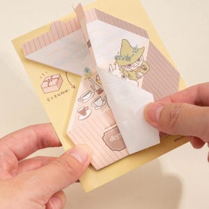 May include: A person is holding a Moomin-themed notepad with a yellow backing. The notepad features illustrations of Moomin characters, coffee cups, and gift boxes. The notepad has a light pink and brown color scheme.