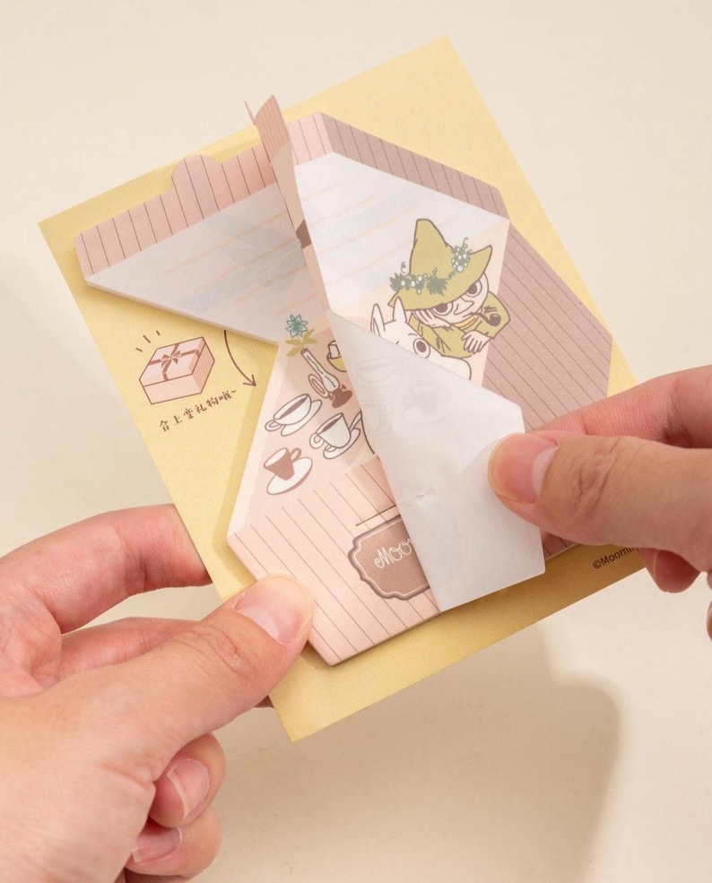 May include: A person is holding a Moomin-themed notepad with a yellow backing. The notepad features illustrations of Moomin characters, coffee cups, and gift boxes. The notepad has a light pink and brown color scheme.