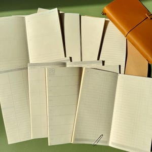 Könnte beinhalten: Eine Sammlung offener Papiernotizbücher und Planer mit verschiedenen Raster-, Linien- und Leerlayouts, cremefarbenen Seiten und einem hellbraunen Ledereinband. Die Seiten zeigen gedruckte Kopfzeilen, Datumsfelder und strukturierte Spalten.
