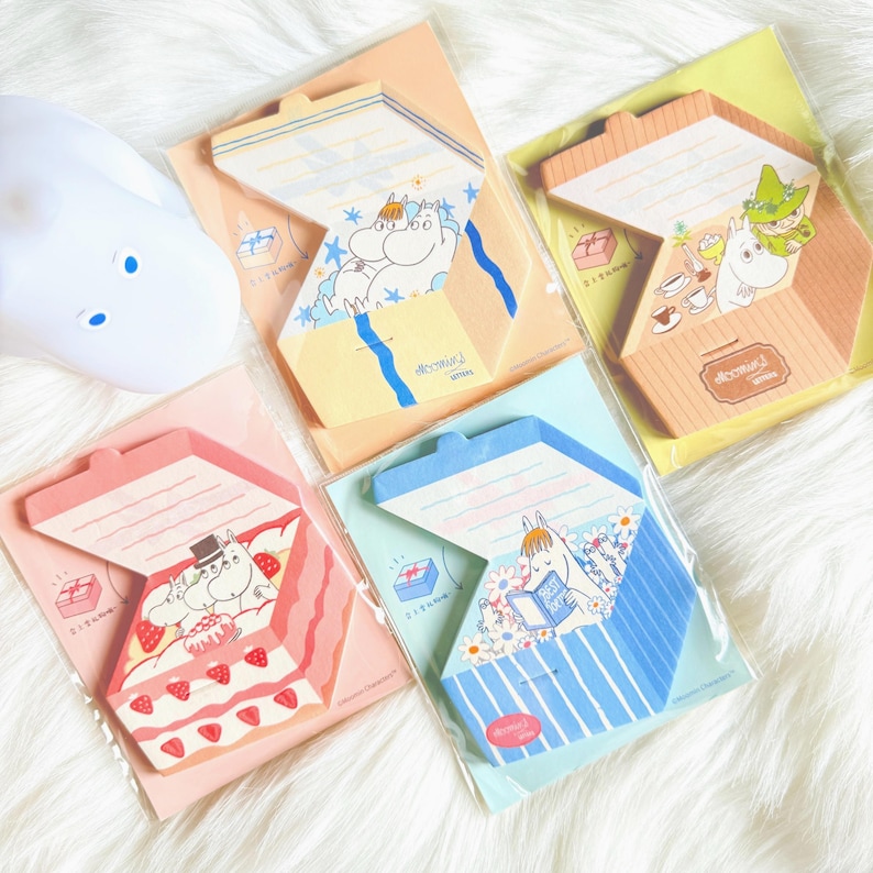 May include: A collection of colorful, illustrated memo pads featuring cartoon characters. Each pad has a unique design, including a cake, a river scene, and a character reading. The pads are packaged in clear plastic.