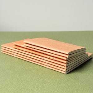 May include: A stack of six rectangular notebooks with light brown, textured paper covers. Each notebook is bound with visible silver staples along the spine, featuring a slim, elongated profile with clean, sharp edges and a consistent, uniform design.