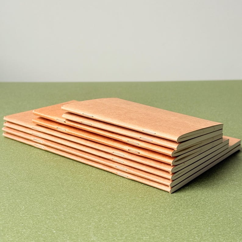 May include: A stack of six rectangular notebooks with light brown, textured paper covers. Each notebook is bound with visible silver staples along the spine, featuring a slim, elongated profile with clean, sharp edges and a consistent, uniform design.