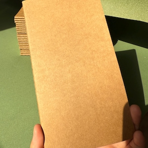 May include: A rectangular notebook with a plain, light brown kraft paper cover. The notebook features a stitched spine visible along the left edge, and it is part of a small stack of identical notebooks, showcasing a simple, functional design.