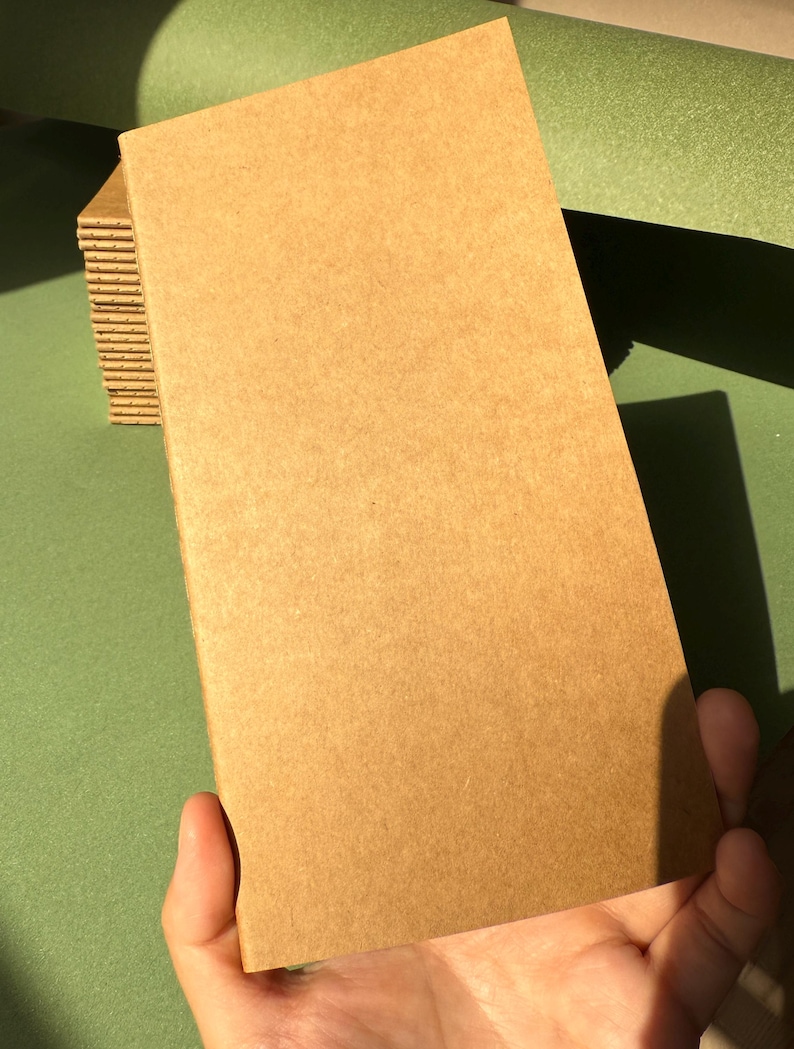 May include: A rectangular notebook with a plain, light brown kraft paper cover. The notebook features a stitched spine visible along the left edge, and it is part of a small stack of identical notebooks, showcasing a simple, functional design.