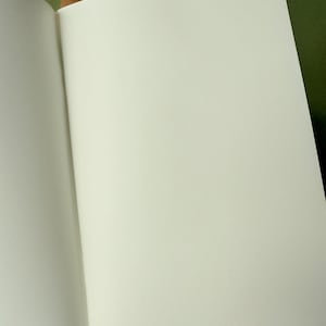 May include: An open notebook with blank, off-white pages and a simple, light brown paper cover. The notebook is held open to reveal the smooth, unlined interior paper, while additional closed notebooks with similar brown covers are stacked behind it.