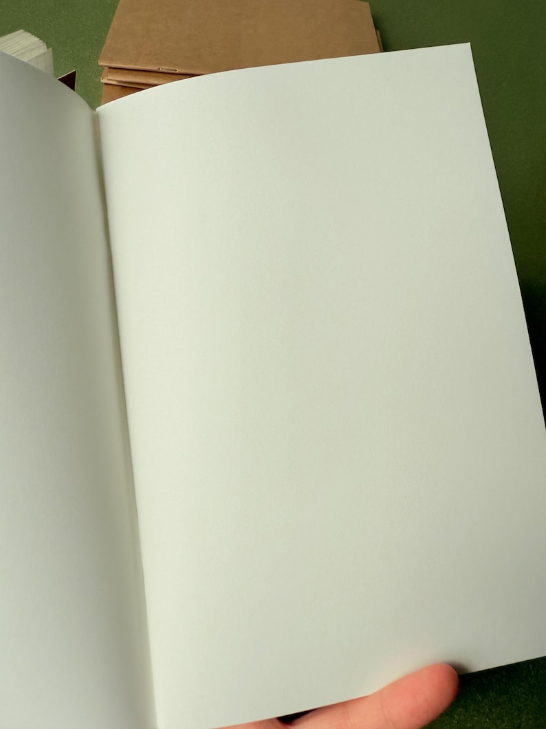May include: An open notebook with blank, off-white pages and a simple, light brown paper cover. The notebook is held open to reveal the smooth, unlined interior paper, while additional closed notebooks with similar brown covers are stacked behind it.