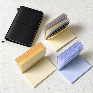 May include: A black leather-bound notebook with an elastic closure strap, accompanied by three open notebooks featuring pages in gradient shades of yellow, orange, purple, and blue, arranged on a textured white surface.