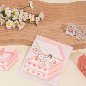 May include: Pink and white Moomin-themed sticky notes shaped like a cake and gift box. The cake-shaped notes feature a strawberry design and Moomin characters. Daisies and ice cubes are in the background.