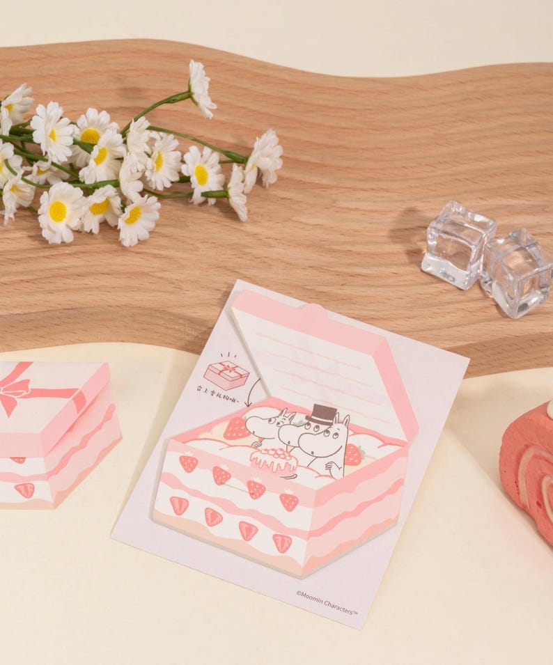 May include: Pink and white Moomin-themed sticky notes shaped like a cake and gift box. The cake-shaped notes feature a strawberry design and Moomin characters. Daisies and ice cubes are in the background.