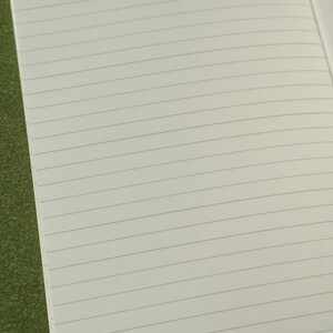 May include: An open notebook with off-white pages featuring thin, horizontal, dark gray lines. The paper has a smooth texture and is arranged in a standard lined format, providing a clean surface for writing or note-taking across the entire page.