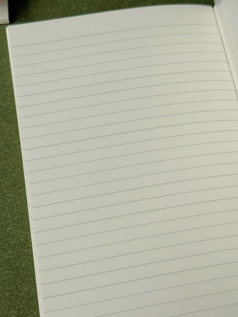 May include: An open notebook with off-white pages featuring thin, horizontal, dark gray lines. The paper has a smooth texture and is arranged in a standard lined format, providing a clean surface for writing or note-taking across the entire page.