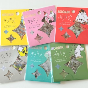 Op de afbeelding: Een verzameling Moomin clear flake seals in geel, roze, rood, zilver, teal en groene verpakkingen. Elk pakket heeft Japanse tekst en een stervormig venster met de stickers. De stickers tonen Moomin-personages.