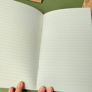 May include: An open notebook with off-white pages featuring thin, horizontal gray lines. The notebook is bound with staples along the center fold. Additional closed notebooks with brown paper covers are visible in the background.