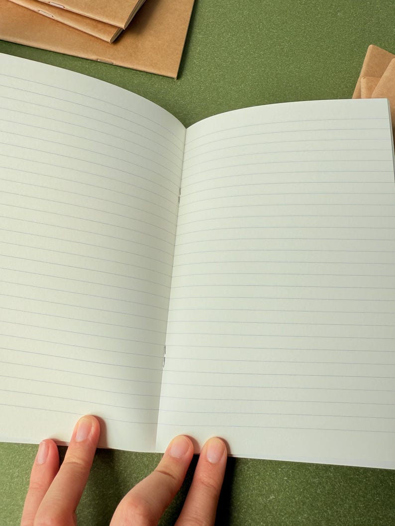 May include: An open notebook with off-white pages featuring thin, horizontal gray lines. The notebook is bound with staples along the center fold. Additional closed notebooks with brown paper covers are visible in the background.