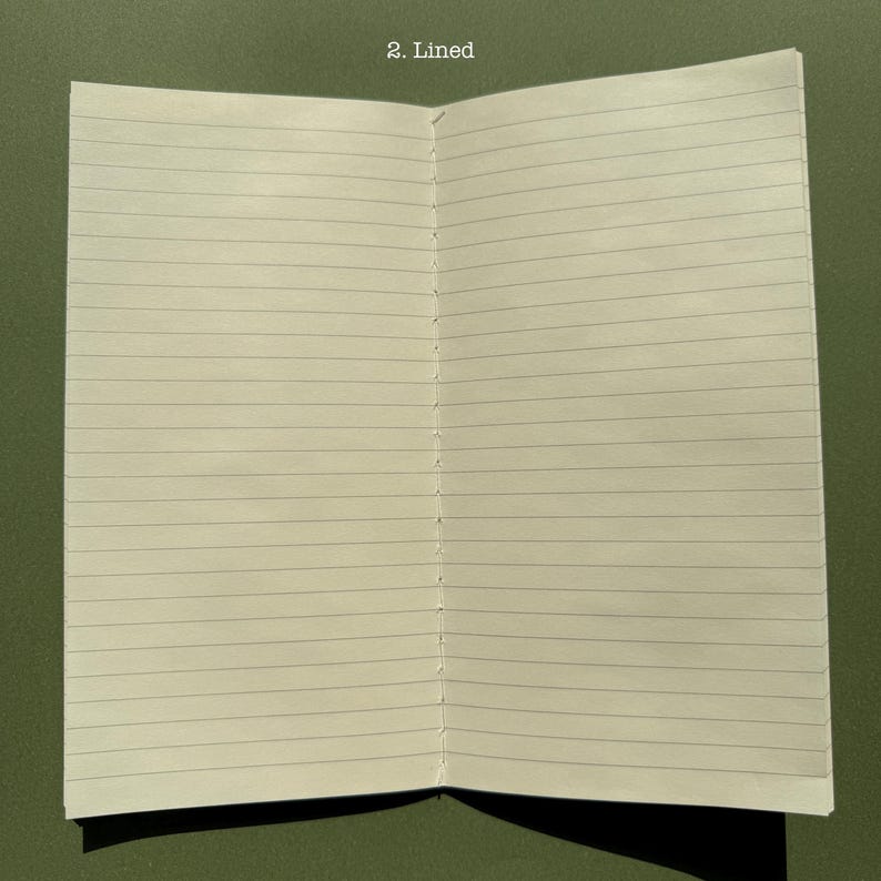 May include: An open notebook with off-white pages featuring horizontal black lines. The center fold shows visible white thread stitching binding the pages together. The text 2. Lined is printed in white at the top center of the page spread.