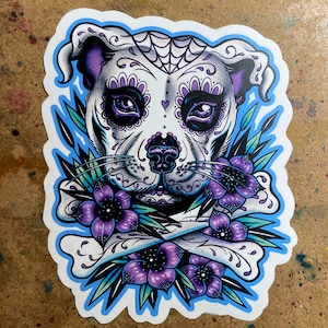 Peut inclure: Un chien en sucre crâne blanc et violet avec une bordure bleue. Le chien est entouré de fleurs violettes et d'os croisés.