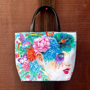 May include: Tote bag with a vibrant illustration of a woman's face, adorned with flowers and cosmic imagery. The design includes a hummingbird, butterflies, and a rainbow. The bag has black handles.