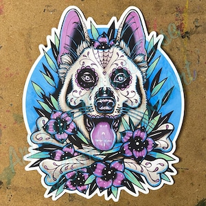 May include: A white German Shepherd with a Day of the Dead sugar skull design, surrounded by purple flowers and crossbones. The dog's tongue is sticking out and the background is blue.