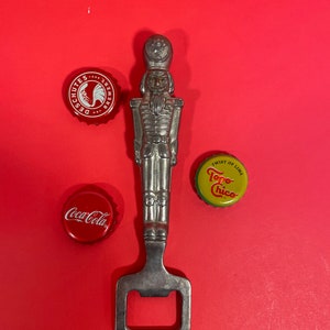 May include: A silver bottle opener shaped like a toy soldier with a red background. The bottle opener is surrounded by three bottle caps: one red with the text "Coca-Cola", one red with the text "Deschutes Brewery", and one yellow with the text "Topo Chico Twist of Lime".