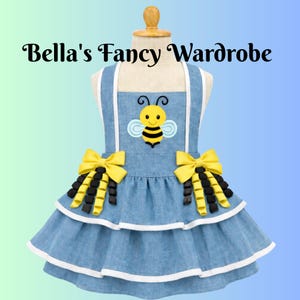 May include: Denim-style pet dress featuring a bee applique and yellow bows. The dress has white trim and the text "Bella's Fancy Wardrobe".