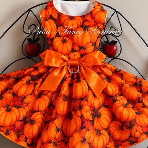 K&ouml;nnte beinhalten: Ein &auml;rmelloses Kleid mit einem Allover-Druck aus orangefarbenen K&uuml;rbissen und Herbstbl&auml;ttern. Das Kleid hat einen Rundhalsausschnitt und eine gro&szlig;e, leuchtend orangefarbene Satinschleife in der Taille mit einem goldfarbenen Ring. Der Text Bella's Fancy Wardrobe ist sichtbar.