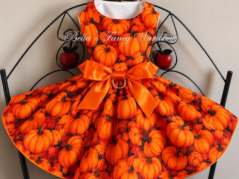 K&ouml;nnte beinhalten: Ein &auml;rmelloses Kleid mit einem Allover-Druck aus orangefarbenen K&uuml;rbissen und Herbstbl&auml;ttern. Das Kleid hat einen Rundhalsausschnitt und eine gro&szlig;e, leuchtend orangefarbene Satinschleife in der Taille mit einem goldfarbenen Ring. Der Text Bella's Fancy Wardrobe ist sichtbar.