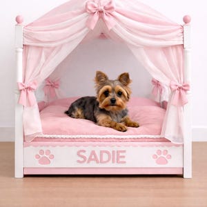 May include: A pink dog bed with a canopy, bows, and the name "Sadie". The bed has a white frame, pink bedding, and a sheer pink canopy. A small dog with brown and black fur is lying on the bed.