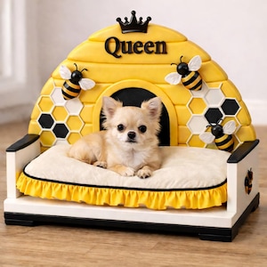 May include: A small, light-colored dog rests on a cream-colored cushion inside a decorative dog bed. The bed is shaped like a yellow beehive with black and white honeycomb details and bee accents. The word "Queen" is displayed on the beehive.